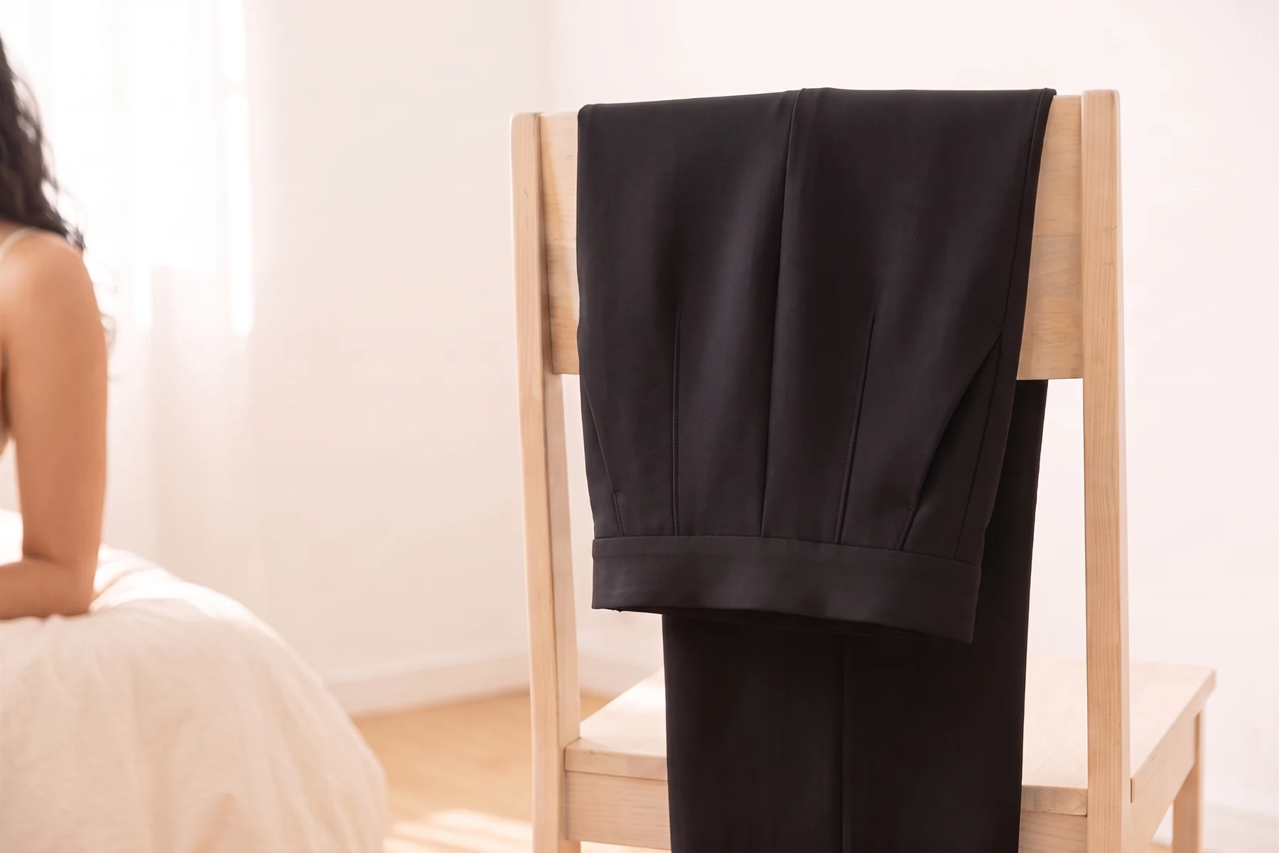 A calça de alfaiataria preta dobrada sobre o encosto de uma cadeira de madeira clara, com o cós alto e um bolso lateral à mostra.