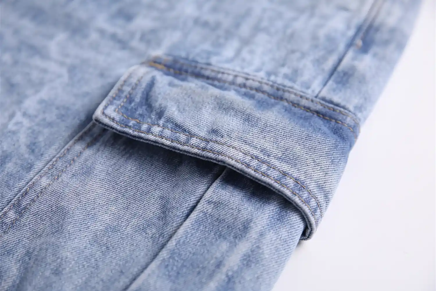 Close do bolso com aba na coxa da calça cargo jeans clara, com o pesponto tan em volta.