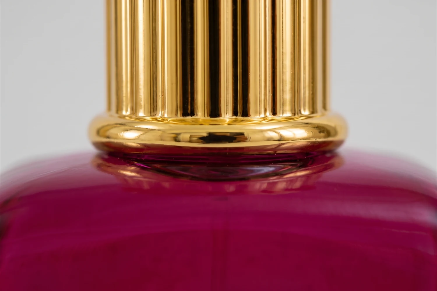 Close do aro dourado canelado da tampa encontrando o ombro magenta do frasco.